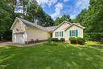 Charming single-story yellow house with green shutters at 95 Wembley Court in Youngsville, featuring an attached two-car garage, a small front porch with chairs, manicured lawn, shrubs, and surrounded by tall trees under a partly cloudy sky.