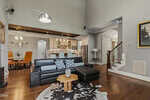 Modern living room with black sectional, wooden coffee table, cowhide rug, and open view into a kitchen with white cabinets and pendant lights. A dining area with orange chairs and decorative deer head is visible on the left.