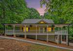 Charming yellow house on 3661 Sandy Creek Road in Franklinton, featuring a large wraparound porch with white railings and hanging plants, surrounded by tall trees at sunset. Warm lights glow from the windows, creating a cozy and inviting atmosphere.