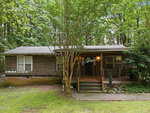 Nestled on a shaded lot at 2658 Goldstone Drive in Oxford, this rustic wooden house features a covered front porch and is surrounded by lush green trees and grass, with a charming stone-lined path leading to the inviting entrance.
