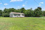 Single-story gray house with white trim and a small front porch, set on a spacious grassy lawn with a gravel driveway at 820 Tungsten Mine Road, Bullock. Surrounded by dense trees and shrubs under a blue sky with scattered clouds.