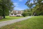 A white two-story house with dormer windows sits on a large green lawn at 2173 Tommie Daniel Road in Oxford. A curved gravel driveway leads to the front of the house, and mature trees surround the property under a blue sky.