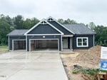 A newly constructed dark blue house with white trim at 65 Kettle Creek Drive, Zebulon, features a triple-car garage, concrete driveway, and unfinished landscaping, surrounded by trees under a cloudy sky.