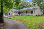 Charming single-story house at 1020 Sagamore Drive in Louisburg, featuring light-colored siding, a gray shingled roof, front porch with white railing, and several steps leading up from a curved driveway. Surrounded by trees and green lawn.