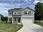 Two-story gray house with white trim, black roof, and attached two-car garage located at 15 Buckthorn Drive, Youngsville. A concrete driveway curves to the garage. The front yard features green grass and small plants near the entrance, with trees and a white fence in the background.