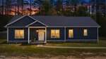 A single-story dark blue house with white trim is shown at dusk at 106 Old NC 75 N in Stem. The porch light is on, illuminating the front door and a chair nearby. Four windows are visible, and there are trees in the background against a colorful sky.