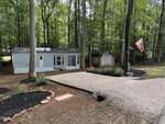 A light blue mobile home sits among tall trees at 113 Kansas Drive, Louisburg, with a gravel driveway, a small shed to the right, a posted mailbox, landscaped flower beds, and a flag on a pole; the area is shaded by a wooded forest.