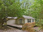 Charming single-story beige house with white trim at 120 Carolina Avenue, Oxford, nestled among dense green trees. The home features a wooden front deck with railings, double glass doors, and several windows. The ground is beautifully landscaped with rocks and shrubs for natural appeal.