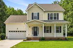 Two-story suburban house with cream-colored siding, dark shutters, a covered front porch supported by columns, and an attached two-car garage. Located at 30 Fern Court in Louisburg, the home features a lush green lawn and is surrounded by trees under a clear blue sky.