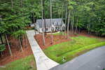 A beige house with a gray roof sits among tall pine trees. A long driveway curves from the road to the front entrance. The lawn is bordered by mulch, and a mailbox is visible at the edge of the driveway near the street.