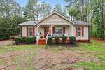 A single-story beige house with a front porch, white trim, and red shutters sits surrounded by trees at 226 Shawnee Drive in Louisburg. Six trimmed bushes line the porch, and a U.S. flag hangs near the front steps. The yard has patches of grass and gravel.