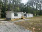 A single-story manufactured home with white siding and a small front porch sits on a gravel driveway at 6669 Brame Drive in Oxford, surrounded by grass and tall pine trees. The sky is partly cloudy.