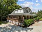 A two-story house at 287 Somerset Lane in Henderson, featuring a gray roof, white siding, large wraparound porch with brown railings, lattice under the porch, surrounded by trees and shrubs, and a concrete driveway in front.