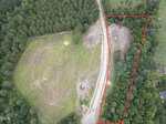 Aerial view of a cleared, grassy area bordered by dense trees at 1 Hidden Dreams Way, Zebulon. A dirt road curves through the scene. A section on the right, outlined in red, includes additional cleared land and forest. A small vehicle is parked near the road.