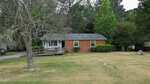 Charming single-story red brick house at 204 Drew Street, Louisburg, featuring a gray roof, two front windows with bushes beneath them, a wheelchair ramp leading to the porch, and a large front yard with patchy grass and several trees.