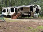 A large Bighorn fifth-wheel RV with a set of wooden stairs sits parked on a gravel and dirt area surrounded by tall trees in a forested campsite at 103 Creek Drive, Louisburg.