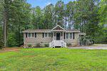 Single-story house with beige siding, white trim, and a small front porch sits on a grassy lawn with gravel driveway at 1379 Briggs Road in Henderson, surrounded by tall pine trees and dense greenery in the background.