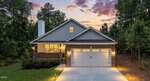 A single-story house at 111 Nashua Drive, Louisburg, features a front porch, brick and gray siding exterior, two-car garage, and lit pathway, surrounded by trees at sunset with a purple and orange sky.