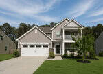 This charming two-story suburban house at 55 Mallory Oak Drive in Franklinton features gray siding, white trim, and a spacious double garage. Enjoy relaxing on the covered front porch or the small balcony above. The landscaped yard includes a lush lawn and a mature tree, while the sky overhead is clear with light clouds.