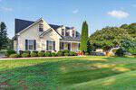 A two-story suburban house with light yellow siding, dark roof, dormer windows, and a covered porch sits gracefully at 90 Torrington Avenue in Franklinton. Manicured bushes line the front, and a large green lawn and tree are visible in the yard under a blue sky.