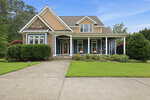 A two-story suburban house at 1530 Raven Wood Drive in Creedmoor featuring blue siding, tan shingle accents, stone detailing, and a front porch with white columns. A concrete driveway leads to the front entrance, which is flanked by green landscaping and two rocking chairs.