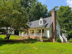 A charming white house with a brick chimney sits on a grassy lawn at 5923 Rivers Bend Drive, Rougemont, surrounded by trees. The house features a front porch with plants and steps, while a detached carport is visible in the background to the left.