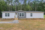 Single-story manufactured home at 6669 Brame Drive in Oxford, featuring white siding, gray accent panels in the center, a small front stoop with stairs, and four windows, set in a grassy clearing with tall trees in the background.