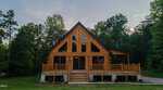 A log cabin with large triangular windows sits in front of a wooded area at 186 Buoy Drive, Henderson. The cabin has a front porch with railings, stairs leading to the entrance, and a peaked roof. The sky is clear with trees on both sides.