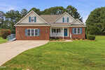 Charming single-story brick house at 40 Perry Lane, Zebulon, featuring white trim, blue shutters, and a blue front door. The home offers a covered front porch, gabled roof, concrete driveway, and is surrounded by a large, neatly mowed lawn with trees in the background.