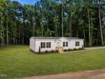 Single-story white manufactured home with a small front porch, surrounded by a large grassy yard and bordered by tall trees in the background. Located at 2558 Buckingham Drive in Franklinton, this charming property features a dirt driveway running along the right side of the house.
