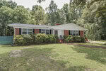 Charming single-story red brick house with white trim, black shutters, and a striking red front door, located at 208 Drew Street in Louisburg. Bushes line the front of the house, and mature trees and grass create a welcoming yard. The home features a classic gable roof and several windows that let in plenty of natural light.