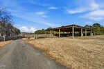 A paved road, possibly 0 Hight Road near Oxford, leads to an open-sided wooden structure with a slanted roof in a dry, grassy field. Trees border the background under a blue sky with scattered clouds.