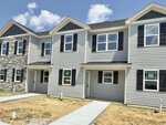 A row of newly constructed two-story townhouses on 1202 Brookrun Drive in Oxford features gray siding, black shutters, and white trim. The front yards are dirt and unfinished, with concrete walkways leading to each entrance. The sky is partly cloudy.