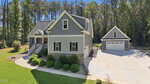 Gray two-story house with stone accents, front porch, and red door, situated on a landscaped yard with shrubs. Separate two-car garage in the background, surrounded by trees and a large concrete driveway.