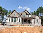 A two-story modern house under construction at 40 Cherry Bark Drive in Youngsville, featuring gray stone and white siding, multiple gables, and a covered front porch. Construction materials and scaffolding are visible, with a dirt yard in the foreground and trees in the background.