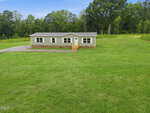 Charming single-story gray manufactured home with white trim and a small front porch, nestled on a large green lawn at 184 Woodsworth Road in Henderson. The property is surrounded by trees and woods in the background under a beautiful blue sky.