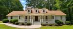 Single-story house with a tan roof, three dormer windows, white brick exterior, covered front porch with white railings, and steps leading to a dark front door. Located at 205 Edgewood Drive in Louisburg, this charming home is surrounded by bushes, grass, and tall trees.