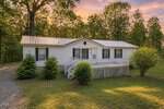 Single-story white house with a metal roof, front porch, ramp, and lattice skirting at 870 Jackson Royster Road in Henderson, surrounded by grass, bushes, and tall trees under a sunset sky. A gravel driveway curves along the left side of the house.