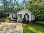 A single-story white house with dark shutters and a gabled roof stands among tall trees on 2764 Gregory Court in Creedmoor. The front yard has trimmed bushes, flowerpots, and a concrete driveway leading to the entrance steps.