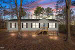 Charming single-story house at 280 Rough Lane, Louisburg, with beige siding, black shutters, and a small wooden front porch. The yard is covered in fallen leaves, while trees surround the property. The sky above glows with colorful sunset hues.