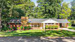 Single-story brick house with large front windows, a central chimney, a covered carport on the right, and well-maintained landscaping in front. Located at 102 Forest Road, Oxford, the home is surrounded by shade trees and sunlight highlights the lush green lawn.