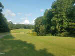 A grassy golf course in Louisburg stretches into the distance, with a curving path on the left and dense, tall green trees lining the right side under a partly cloudy sky near 249 Sacred Fire Road.
