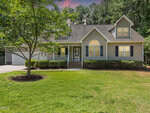 A two-story suburban house with beige siding, black shutters, and a front porch, located at 25 Shellcross Court in Youngsville. The yard is grassy with a tree and trimmed bushes. The driveway leads to a two-car garage, while tall trees line the background.