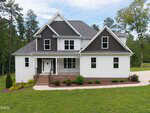 Two-story modern house with white siding and dark gray gables, located at 190 Whistlers Cove, Louisburg. Brick steps lead to a covered front porch with black railing. The home is surrounded by grass, shrubs, and trees with a driveway on the right.