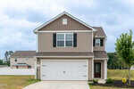 Two-story beige house with white trim, black shutters, and a double garage door at 106 Long Green Way in Oxford. A concrete driveway leads to the entrance, and a small green lawn surrounds the home. The sky is cloudy and other homes are visible in the background.
