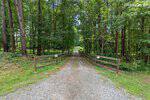 A gravel driveway flanked by wooden fences leads through a dense, green forest. Signs are posted on both sides of the entrance, and the scene is well-lit by natural daylight.
