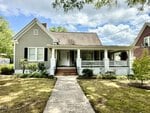 A light gray house with white trim and black shutters sits gracefully on 106 High Street in Oxford, featuring a large front porch with white railings. Brick steps lead to the front door, while the yard includes small bushes and trees, complemented by a concrete walkway to the inviting porch.