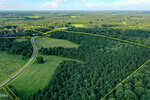 Aerial view of a rural landscape along 38 Acres Timberlake Road, Louisburg, with a winding road, grassy fields, dense forest, and property boundaries marked by yellow lines. A single red vehicle is visible on the road beneath a partly cloudy sky.