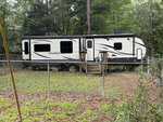 A white and black travel trailer sits behind a chain-link fence in a grassy, wooded yard at 108 Poteau Drive in Louisburg. There are wooden steps leading to the door, and trees surround the area.