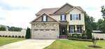 Two-story suburban house at 15 Brushwood Court, Youngsville, with tan siding, stone accents, a double garage, and a manicured front yard featuring shrubs and a small flower bed. The driveway leads to the garage, and a white fence encloses the backyard.